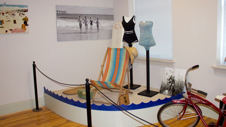 The beach gallery: period swimsuits on stands and a striped deck chair on a low blue-edged platform, a red bicycle to one side and a large black and white beach photograph on the wall.