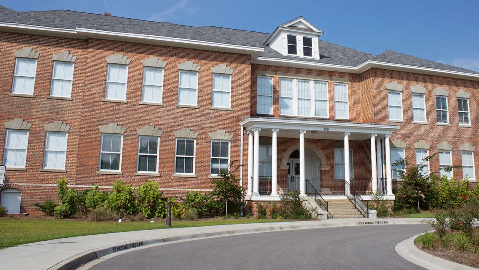 The museum building today, seen from the entrance loop: a symmetrical two-story red brick school building with a white portico, dormer and clipped shrubs.