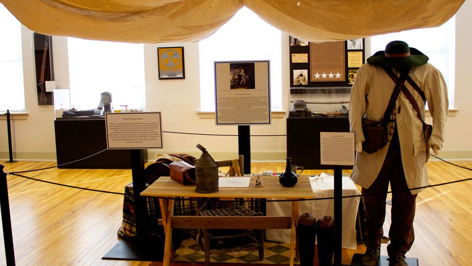 The military gallery: a canvas campaign tent over a table of camp equipment, with a figure in a long coat and broad hat standing beside it and framed documents behind.