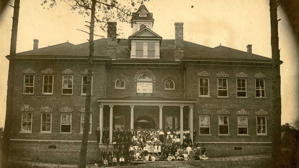 A sepia photograph of the Burroughs School soon after it was built, with a large group of pupils and teachers assembled across the front steps.