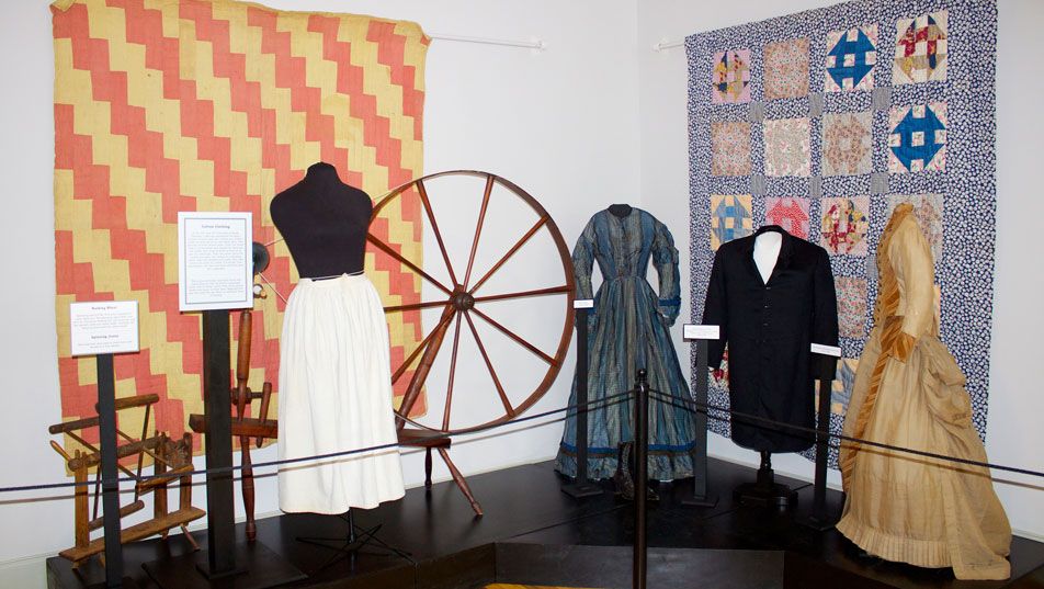 The textiles gallery: a coral and cream zigzag quilt and a blue block quilt hung on the walls, with a spinning wheel and four dressed mannequins in period clothing between them.