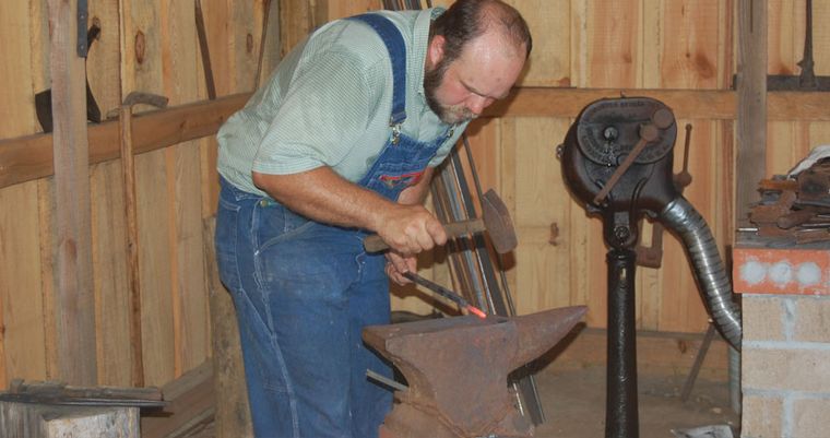A blacksmith in overalls works a length of iron over the anvil in the timber-framed blacksmith shop.