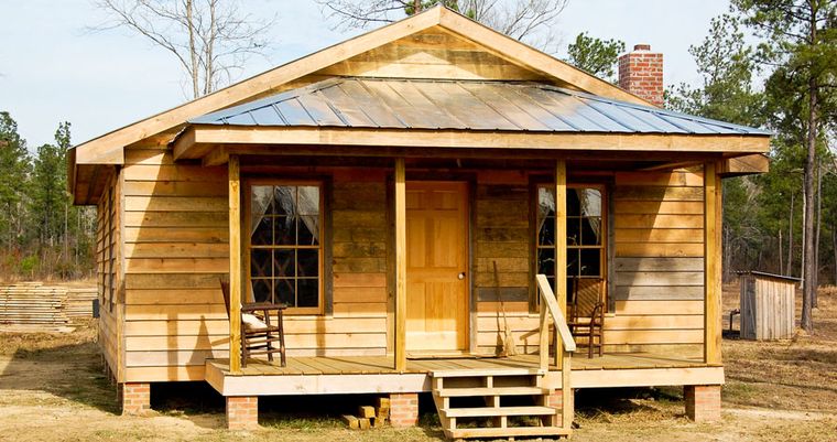 A board-and-batten wooden house at the L.W. Paul Living History Farm, set on brick piers under a metal roof, with rocking chairs on its front porch and wooden steps down to the yard.