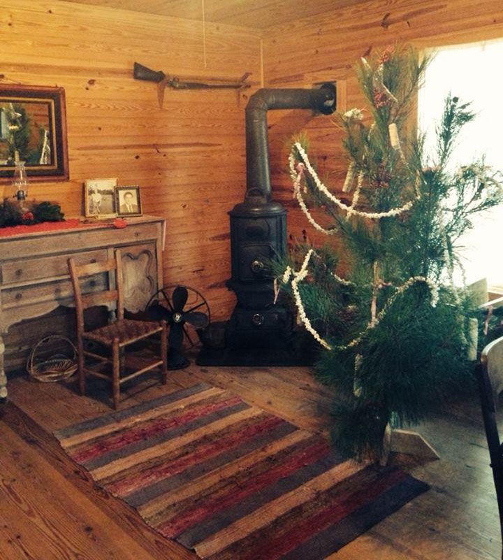 The farmhouse front room at Christmas: a lit tree beside a cast-iron stove, a rag rug on the plank floor and a rocking chair by the wall.