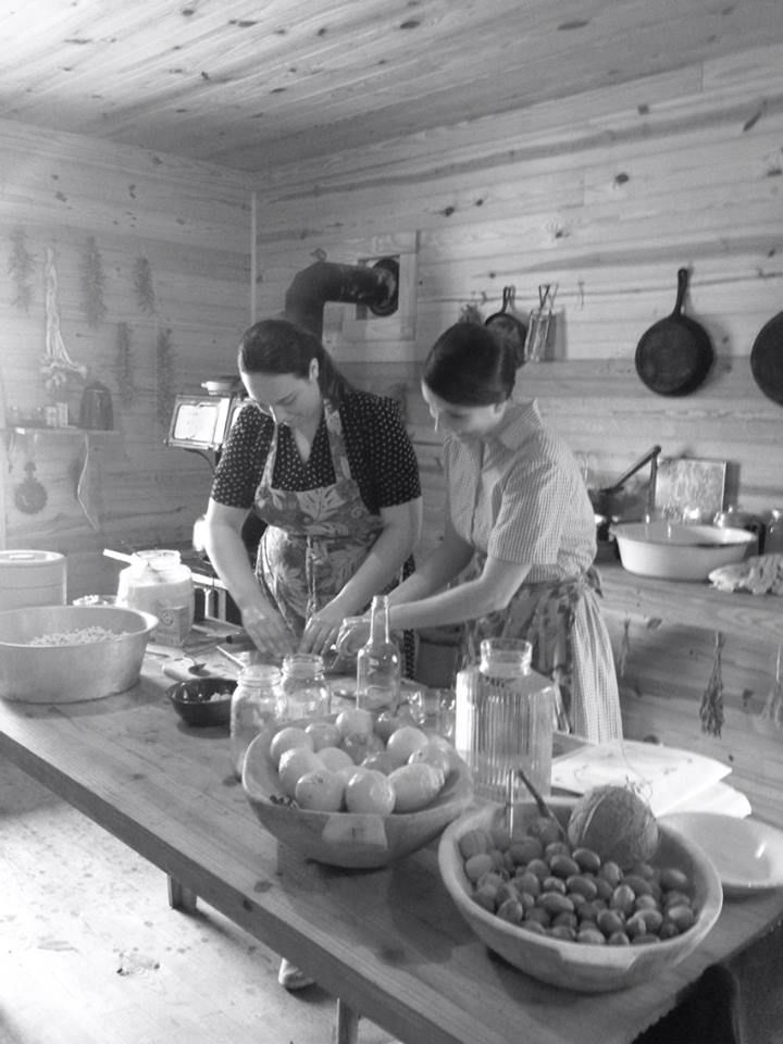 Black and white photograph of two women in aprons preparing food at the farmhouse kitchen table, bowls of fruit and vegetables in front of them.