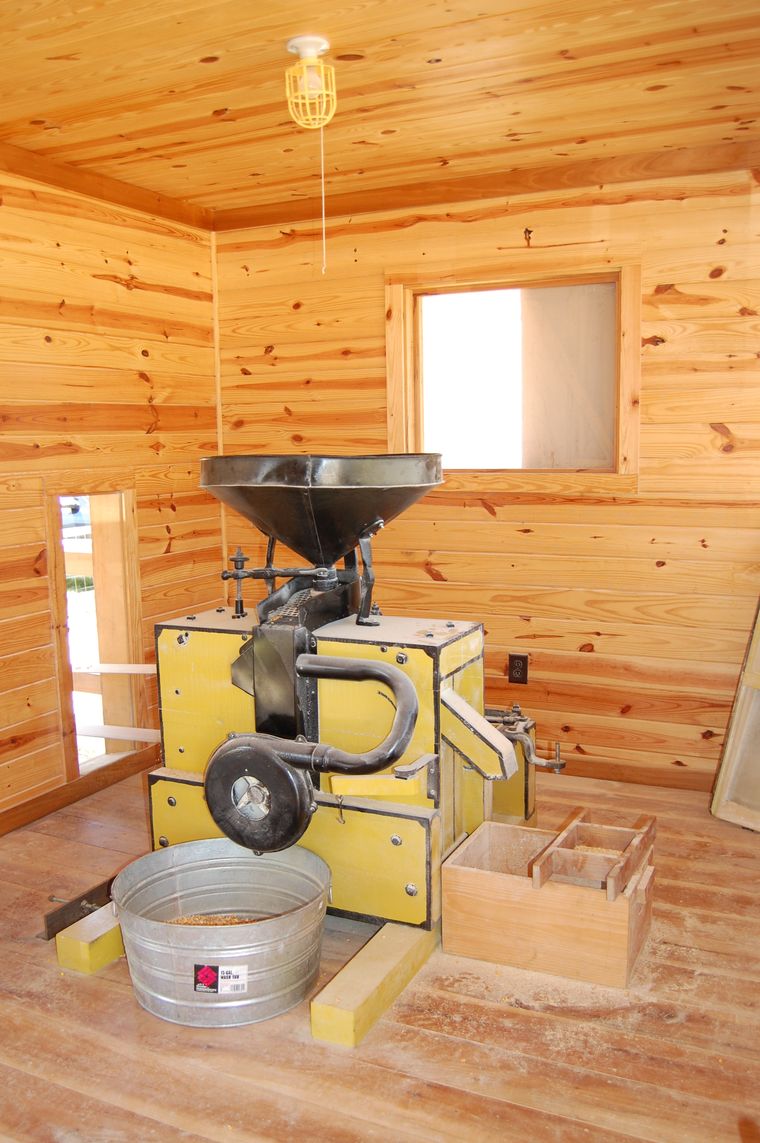 The gristmill interior: a yellow belt-driven milling machine and hopper against the bright pine boards of the mill house.
