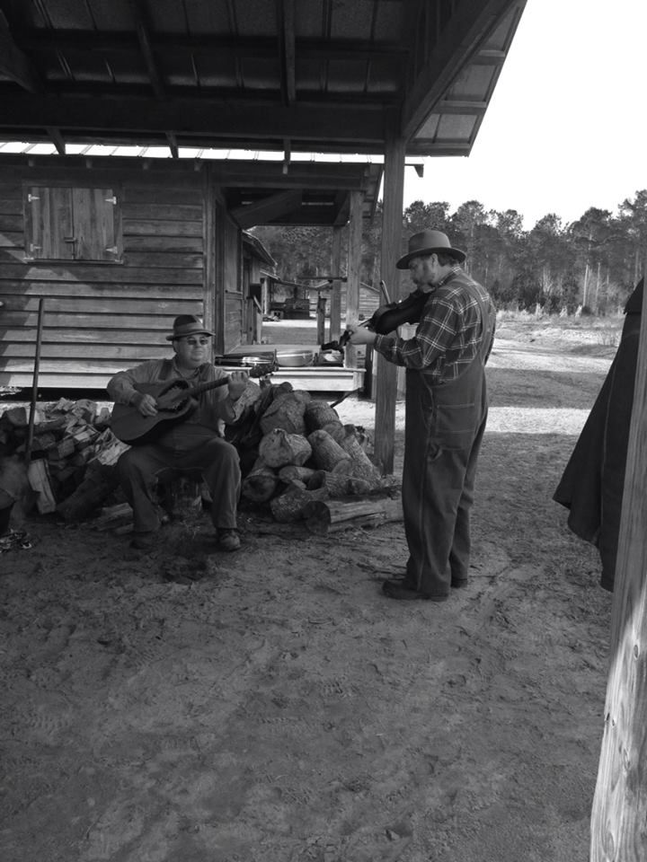 Black and white photograph of two men in overalls and hats making music under the shelter of an outbuilding, one seated with an instrument.
