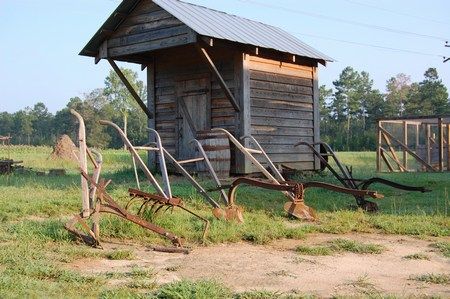 The smokehouse, a small board-and-batten building with a metal roof, with several horse-drawn plows lined up in the grass in front of it.