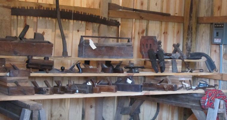 The carpenter shop wall, hung with hand saws, planes, augers and braces above a cluttered workbench.