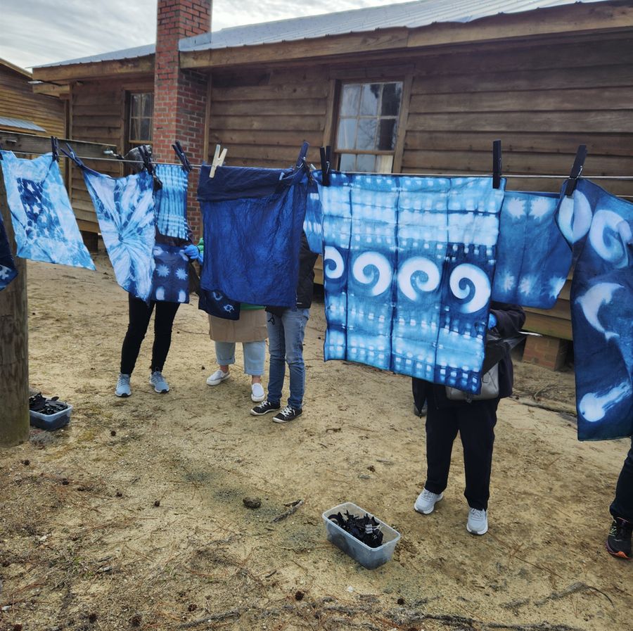 Indigo-dyed cloths in blues and whites pegged on a line outside the farm buildings, three people holding them up to show the patterns.
