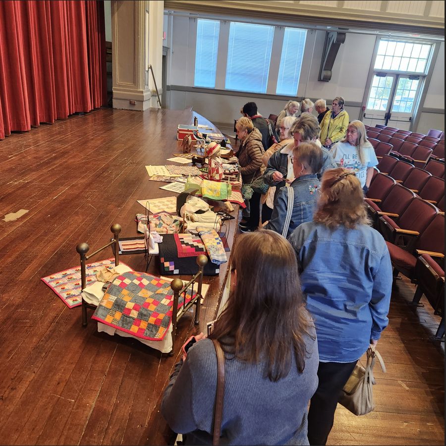 Visitors filing along a row of tables laid with quilts in the auditorium, tall windows down one side.
