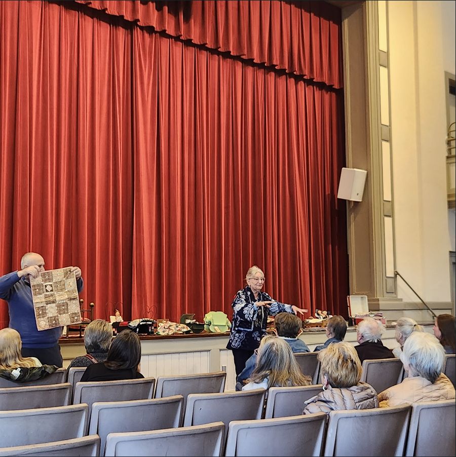 A presenter on the McCown Auditorium stage holding up a quilt in front of a seated audience, the red stage curtain behind.