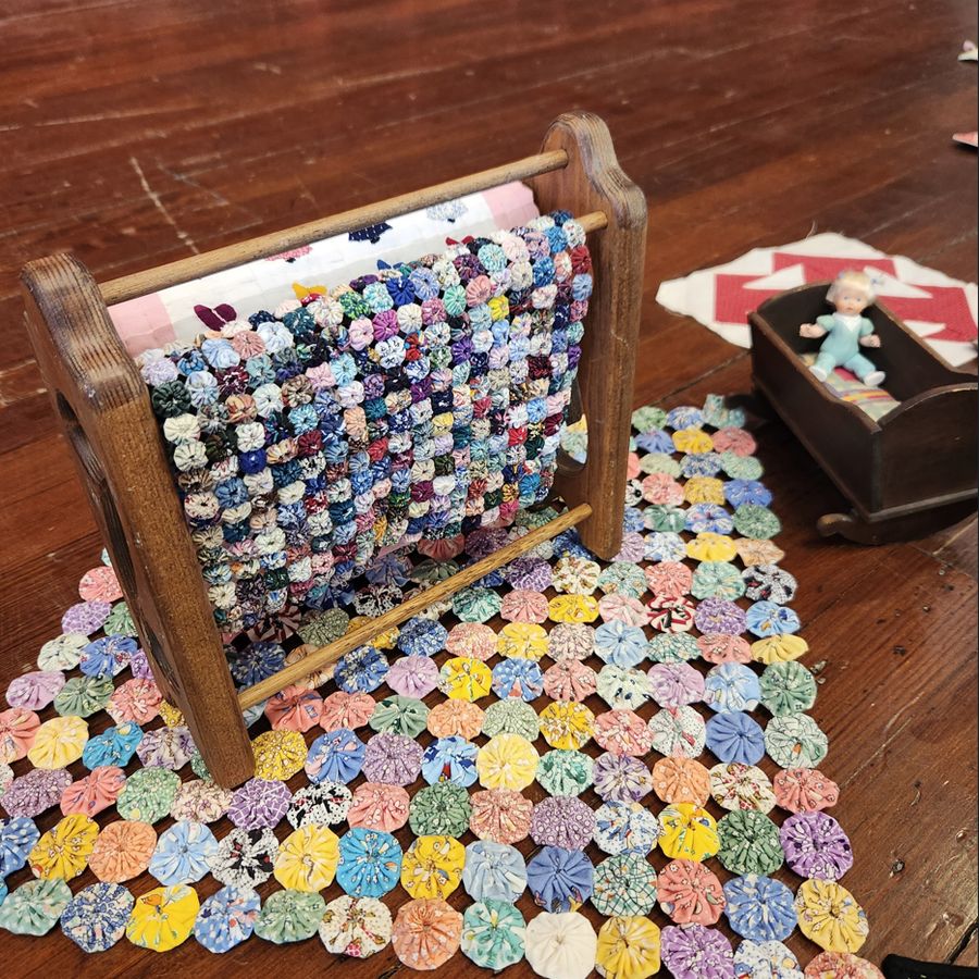 A quilt of hand-stitched fabric yo-yos spread across a plank floor, part of it still stretched on a wooden quilting frame.